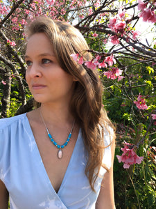 Vintage pink coral pendant hangs from turquoise bead necklace and handmade silver bead necklace on woman in blue dress with pink cherry blossom trees in background